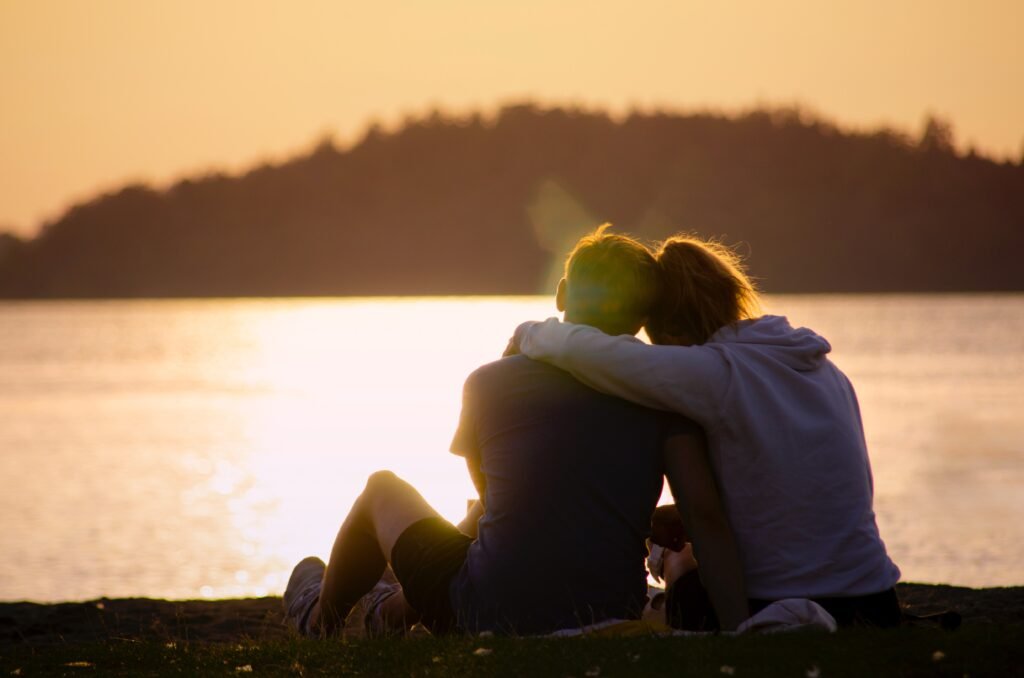 a couple (a girl and a boy) are facing a body of water. The girl has her arm around the shoulder of the boy.